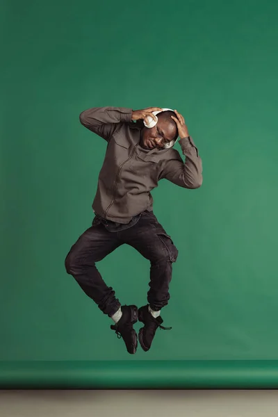 Studyjne ujęcie afrykańskiego mężczyzny w słuchawkach skaczących i słuchających muzyki odizolowanych na ciemnozielonym tle — Zdjęcie stockowe
