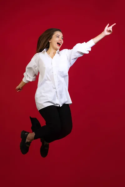 Joven optimista hermosa niña con camisa blanca riendo y saltando aislado sobre fondo de estudio de color rojo oscuro. Concepto de emociones, moda — Foto de Stock