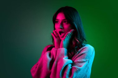 Stüdyo çekimlerinde sevimli bir kız, günlük kıyafetleri içinde koyu yeşil bir stüdyoda izole edilmiş pembe neon ışığı altında poz veriyor..