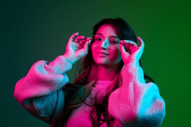 Stüdyo çekimlerinde sevimli bir kız, günlük kıyafetleri içinde koyu yeşil bir stüdyoda izole edilmiş pembe neon ışığı altında poz veriyor..