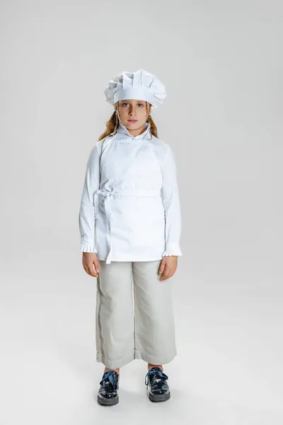 Retrato de niña linda en uniforme de cocinero blanco y enorme sombrero de chefs de pie aislado sobre fondo de estudio blanco. — Foto de Stock