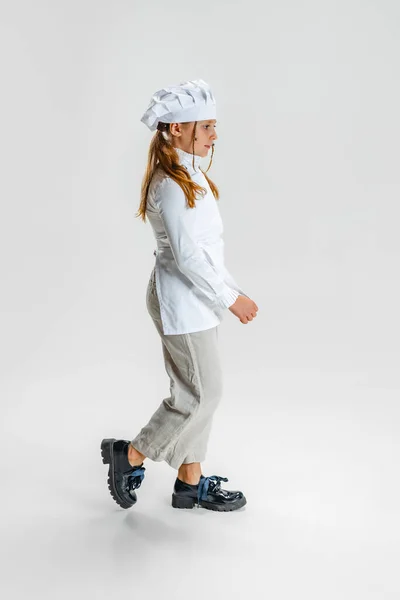 Liten vit flicka i vit kock uniform och stora kockar hatt går isolerad på vit studio bakgrund. — Stockfoto
