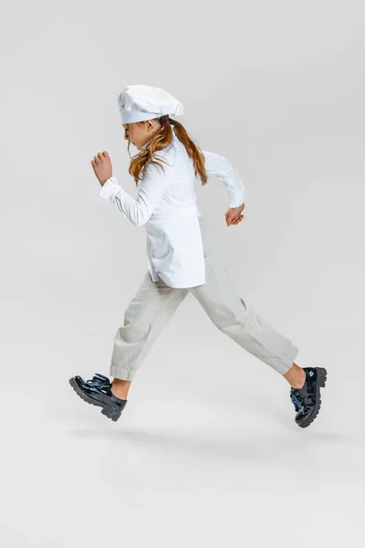 Liten vit flicka i vit kock uniform och stora kockar hatt kör isolerad på vit studio bakgrund. — Stockfoto