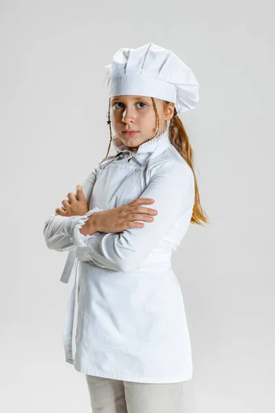 Retrato de media longitud de la niña de la escuela en uniforme de cocinero blanco y enorme sombrero de chefs de pie con los brazos cruzados aislados en el fondo del estudio blanco. —  Fotos de Stock