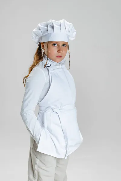 Halblanges Porträt eines kleinen Schulmädchens, einer Studentin in weißer Kochuniform und einem riesigen Kochmütze, die isoliert auf weißem Studiohintergrund posiert. — Stockfoto