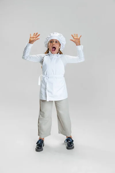 Retrato de niña linda en uniforme de cocinero blanco y sombrero de chefs enormes levanta las manos aisladas sobre fondo de estudio blanco. — Foto de Stock