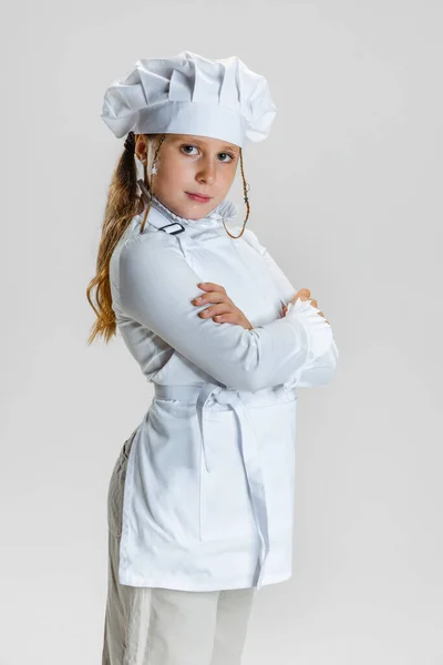 Halblanges Porträt eines kleinen Schulmädchens in weißer Kochuniform und riesigem Kochmütze, das isoliert mit verschränkten Armen auf weißem Studiohintergrund steht. — Stockfoto