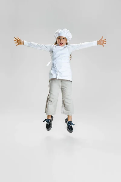 흰색 요리 유니폼을 입고 화이트 스튜디오 배경에서 분리 된 대형 요리사 모자를 쓰고 있는 귀여운 여자 아이의 다이나믹 이미지. — 스톡 사진