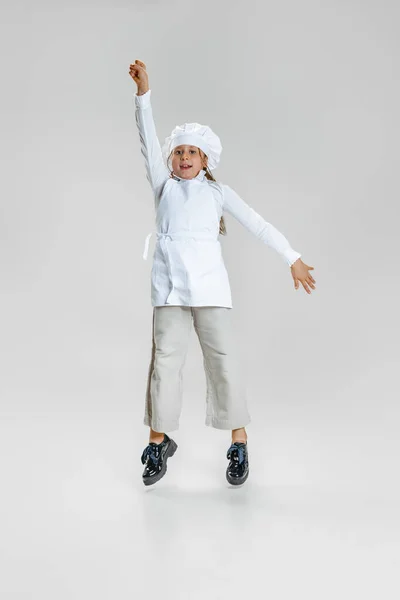Imagen dinámica de la niña linda en uniforme de cocinero blanco y enormes chefs sombrero saltando aislado sobre fondo de estudio blanco. — Foto de Stock