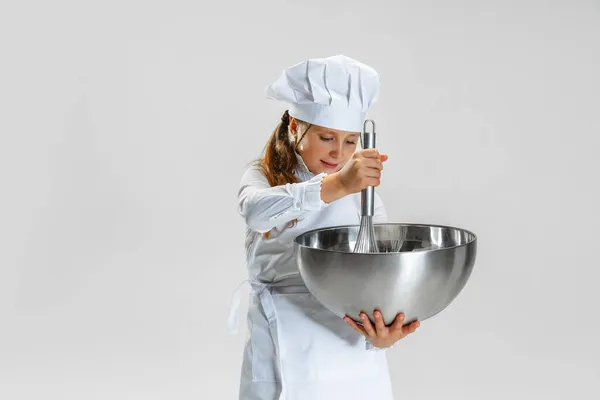 Ein kleines nettes Mädchen in weißer Kochuniform und riesigem Kochmütze posiert isoliert auf weißem Studiohintergrund. — Stockfoto