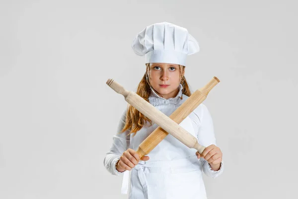 Uma menina fofa no uniforme cozinheiro branco e enorme chapéu de chefs posando isolado no fundo do estúdio branco. — Fotografia de Stock