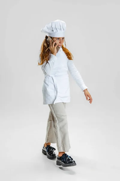 Full längd porträtt av liten flicka i vit kock uniform och stora kockar hatt poserar isolerad på vit studio bakgrund. — Stockfoto