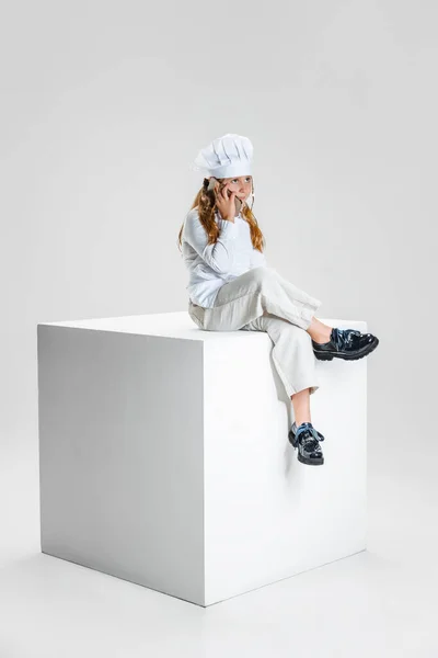 Fényképezés a kislány, diák fehér szakács egyenruha és hatalmas szakácsok kalap ül hatalmas doboz elszigetelt fehér stúdió háttér. — Stock Fotó