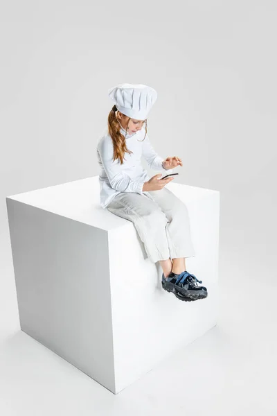 Fotografering av liten flicka, student i vit kock uniform och stora kockar hatt sitter på stor låda isolerad på vit studio bakgrund. — Stockfoto