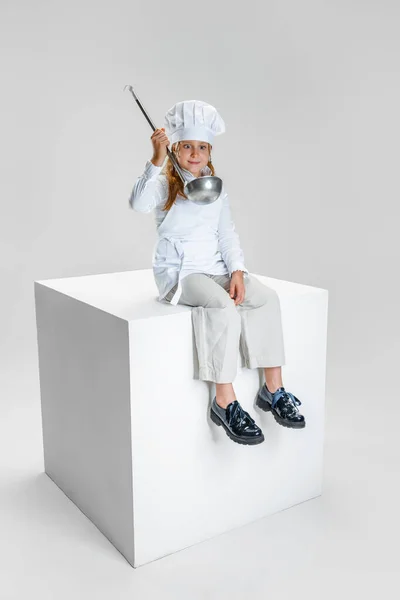 Fotografie holčičky, studentky v bílé kuchařské uniformě a obrovský kuchařský klobouk sedí na obrovské krabici izolované na bílém pozadí studia. — Stock fotografie