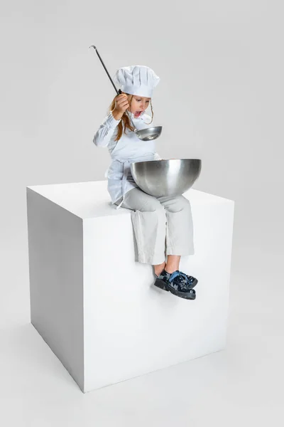 Fotografía de niña, estudiante en uniforme de cocinero blanco y sombrero de chefs enormes sentados en una caja enorme aislada sobre fondo de estudio blanco. —  Fotos de Stock