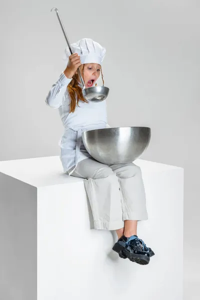 백인 요리사 복장을 한 학생 과 거대 한 요리사 모자를 쓴 작은 소녀의 사진 이 흰색 스튜디오 배경 위에 고립된 거대 한 상자 위에 앉아 있는 모습. — 스톡 사진