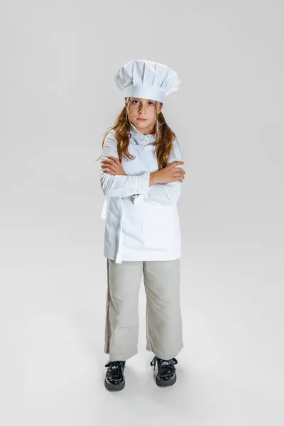 Retrato de larga duración de niña en uniforme de cocinero blanco y sombrero de chefs enormes posando aislado sobre fondo de estudio blanco. — Foto de Stock