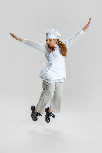 Dynamisk bild av liten söt flicka i vit kock uniform och stora kockar hatt flyger isolerad på vit studio bakgrund. — Stockfoto
