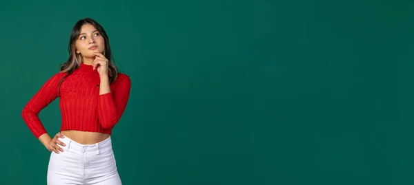 Horizontal flyer with young beuatiful girl, student in bright red sweater thinking isolated on green studio backgroud. — Stock Photo, Image