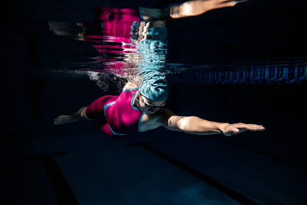 En kvinnlig simmare i badmössa och skyddsglasögon tränar vid poolen, inomhus. Undervattensvy över simning detaljer. — Stockfoto