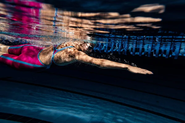 Egy női úszó úszósapkában és védőszemüvegben edz a medencében, bent. Vízalatti kilátás az úszási mozgásokra részletek. — Stock Fotó