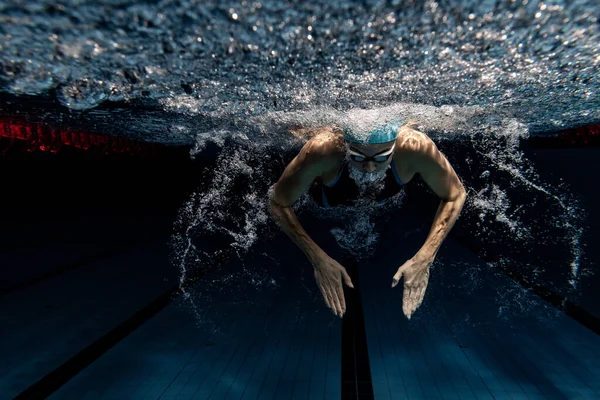 Vista submarina de los detalles de los movimientos de natación. Una nadadora en gorra de natación y gafas de entrenamiento en la piscina, en interiores. — Foto de Stock