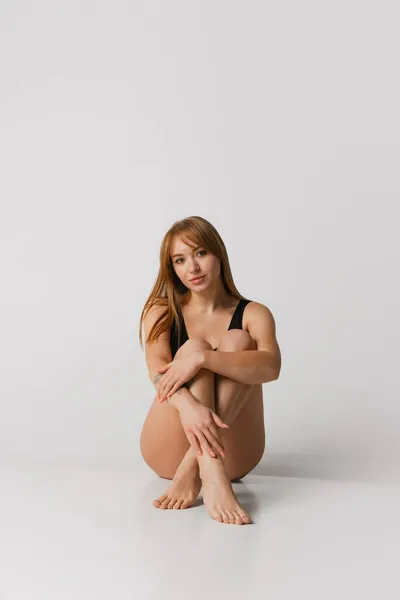 Jovem linda menina ruiva em maiô preto sentado no chão isolado sobre fundo estúdio cinza. Conceito de beleza natural. — Fotografia de Stock