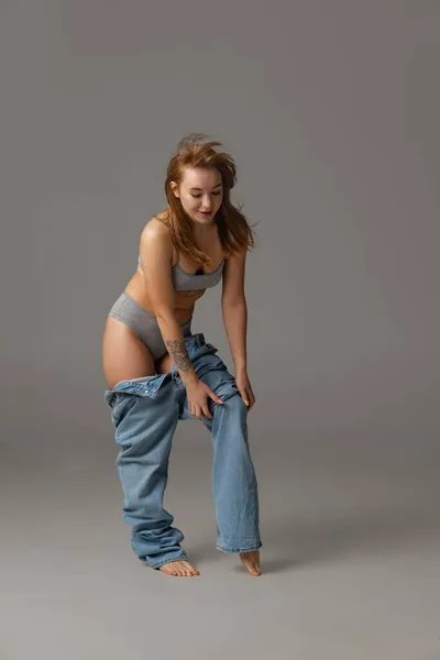 Young beautiful red-headed woman putting blue jeans jumpersuit on isolated over gray studio background. Weight Loss concept. — Stock Photo, Image