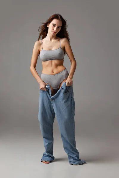 Retrato de joven hermosa chica delgada en pantalones vaqueros azules jumpersuit posando aislado sobre fondo de estudio gris. concepto de pérdida de peso. — Foto de Stock