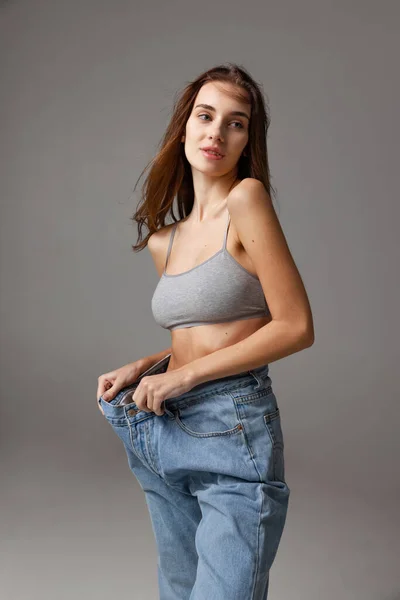 Ritratto di giovane bella ragazza magra in tuta blu jeans posa isolata su sfondo grigio studio. Perdita di peso concetto. — Foto Stock