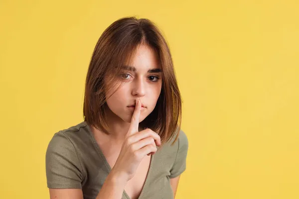 Retrato de chica bonita joven en camiseta marrón sostiene su dedo cerca de los labios aislados en el fondo del estudio amarillo. —  Fotos de Stock