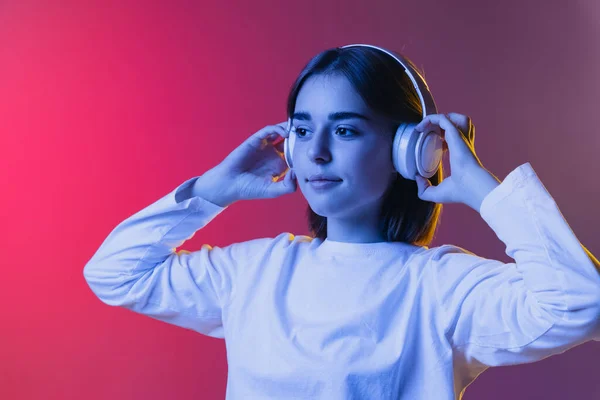 Primo piano ritratto di giovane ragazza carina, studente in abiti casual ascoltare musica isolata su rosa-viola studio backgroud in neon. — Foto Stock