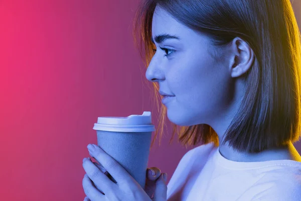 Tampilan samping, profil gadis muda yang lucu, siswa berpakaian kasual minum kopi terisolasi pada merah muda-ungu gradien studio backgroud di neon. — Stok Foto