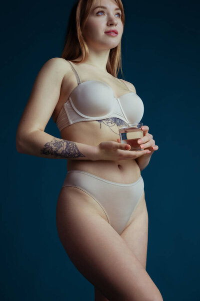 Портрет молодой женщины в обнаженном цвете нижнего белья со стаканом воды изолирован на темно-синем фоне студии. Концепция природной красоты.