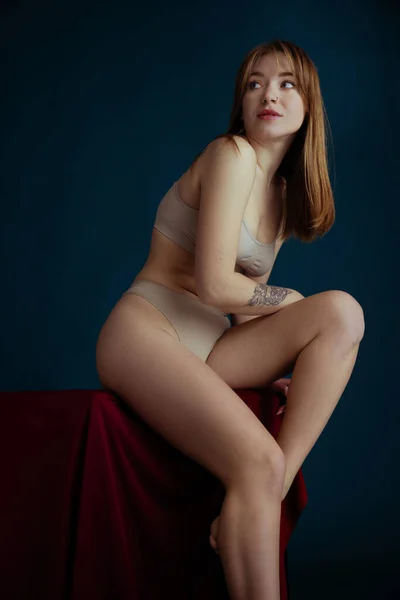 Classico ritratto di giovane bella donna rossa in biancheria intima di colore nudo posa isolata su sfondo blu. — Foto Stock
