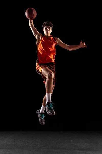 Full length portrait of basketball player training isolated on dark studio background. Tall muscular athlete jumping with ball. — Stock Photo, Image
