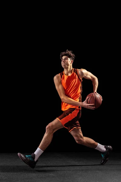 Full length portrait of basketball player training isolated on dark studio background. Tall muscular athlete jumping with ball. — Stock Photo, Image