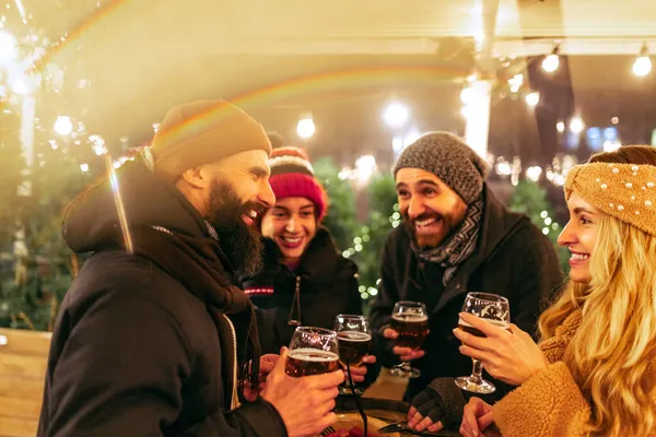 Giovani felici, amici con bicchieri di birra e salsicce ridono, trascorrendo del tempo insieme alla fiera invernale la sera. Vacanze, concetto di Natale — Foto Stock
