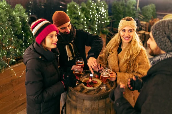 Felices amigos sonrientes con copas de vino caliente que se divierten, pasando tiempo juntos en la feria de invierno por la noche. Vacaciones, concepto de Navidad — Foto de Stock