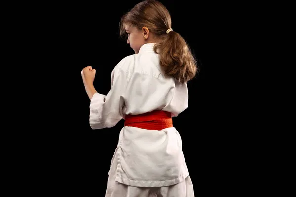 Bakåt syn på liten flicka, ung karate i kampen hållning isolerad över mörk bakgrund. Begreppet idrott, utbildning, färdigheter — Stockfoto