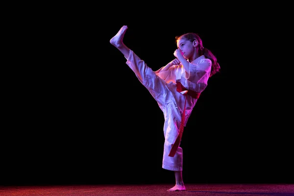 Portret van een klein meisje, jong karate dat alleen oefent, geïsoleerd over een donkere achtergrond. Begrip sport, onderwijs, vaardigheden — Stockfoto
