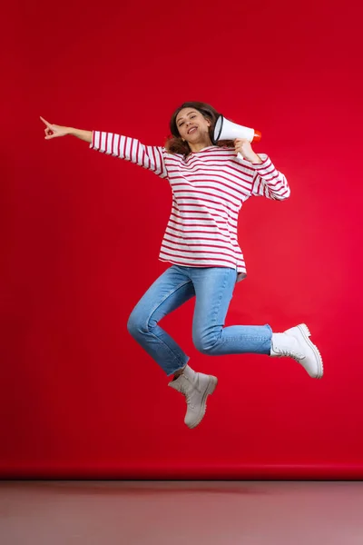 Menina bonita jovem emocional em suéter e jeans pulando isolado no fundo do estúdio vermelho. Conceito de emoções, expressão facial — Fotografia de Stock