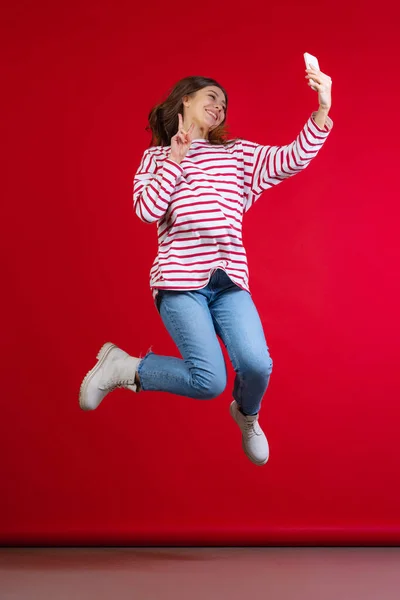 Kırmızı stüdyo arka planında selfie çeken hırkalı ve kot pantolonlu duygusal genç bir kız. Duyguların kavramı, yüz ifadesi — Stok fotoğraf