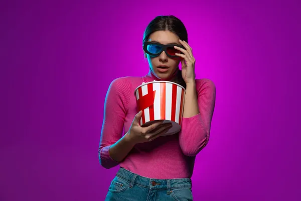 Одна молода красива емоційна дівчина дивиться фільм ізольовано на фіолетовому студійному фоні в рожевому неоновому світлі . — стокове фото