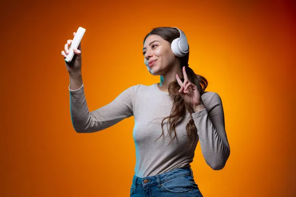 Retrato de meia-comprimento de menina bonita ouvindo música e usando telefone isolado no fundo do estúdio marrom amarelo em luz de néon. — Fotografia de Stock