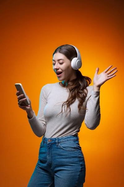 Retrato de meia-comprimento de menina bonita ouvindo música e usando telefone isolado no fundo do estúdio marrom amarelo em luz de néon. — Fotografia de Stock