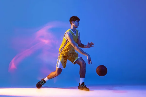 Dynamisch portret van professionele basketbalspeler training met bal geïsoleerd op blauwe studio achtergrond in gemengd neon licht. — Stockfoto