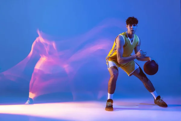 Dynamiczny portret profesjonalnego koszykarza trening z piłką izolowaną na niebieskim tle studio w mieszanym świetle neonowym. — Zdjęcie stockowe