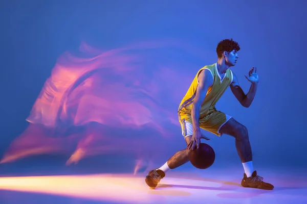 Retrato dinámico de entrenamiento de jugador de baloncesto profesional con pelota aislada sobre fondo de estudio azul en luz de neón mixta. —  Fotos de Stock